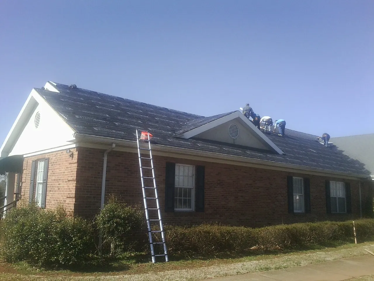 Crew tearing shingles off a brick ranch roof with a ladder set — Metal Roof Repair work by our crew in Brownell