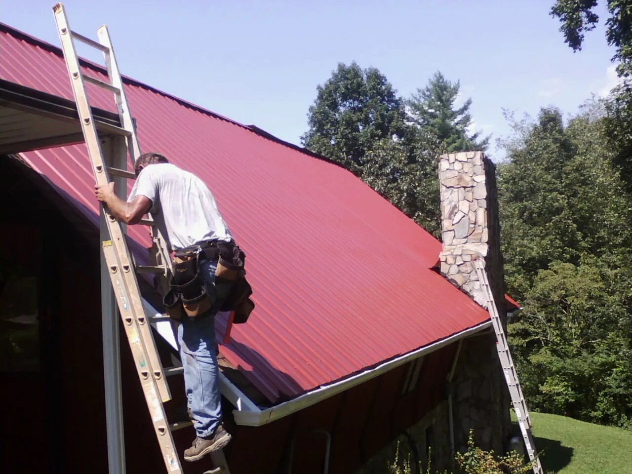 Roofer on a ladder beside a red metal roof and stone chimney — Soffit & Fascia Repair by Roofing Brothers in Brownell